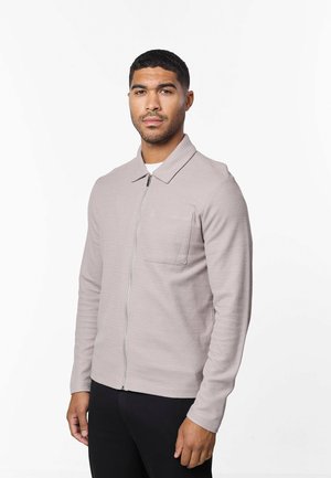 Homme portant une veste zippée gris clair avec un col et une poche poitrine, debout devant un fond blanc uni.