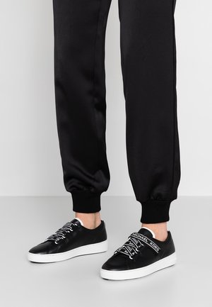 Nogi w czarnych spodniach dresowych i czarnych niskich trampkach z białymi podeszwami i sznurowadłami z napisem „Michael Kors”, stojące na szarej podłodze.