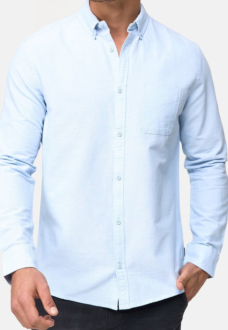 Camisa azul claro de botones, con un bolsillo, mangas largas y una textura suave. Presenta un cuello clásico y cierre frontal de botones.
