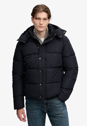 Giovane uomo con giacca imbottita nera con cappuccio e jeans blu, in piedi con una mano in tasca davanti a uno sfondo uniforme.