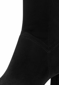 Ghetă de gleznă din piele intoarsă neagră, cu o textură netedă, formă adaptată și toc înalt. Prezintă cusături detaliate de-a lungul cusăturii.