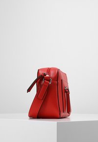 Červená kožená taška se strukturovaným tvarem, má nastavitelné ramenní popruhy, boční kapsu a stříbrné kovové doplňky. Hladká textura.