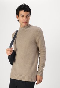Maglione beige con collo alto a costine e dettagli ai polsini. Realizzato in tessuto testurizzato, presenta una vestibilità comoda ed è abbinato a una borsa nera.