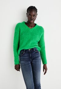 Vihreä pörröinen neule, jossa on V-aukko ja pitkät hihat, yhdistettynä tummansinisiiin skinny-farkkuihin. Neuleessa on teksturoitu pinta.