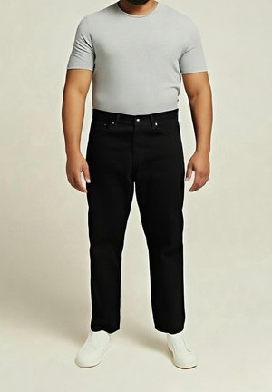Homme portant un t-shirt gris clair ajusté, un pantalon noir et des chaussures blanches sans lacets, debout devant un fond uni.