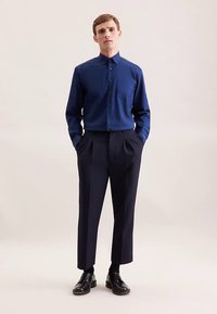 Donkerblauw overhemden met knopen, voorzien van een kraag en een borstzak, gecombineerd met donkerkleurige broek met hoge taille en zwarte leren schoenen. Gladde stoftextuur.