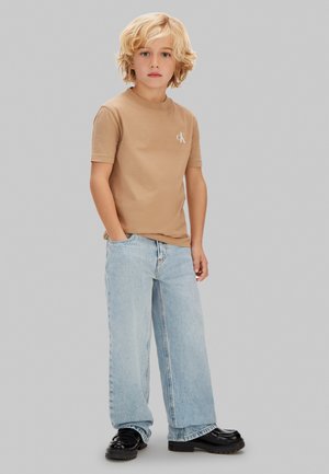 Niño rubio de pie con las manos en los bolsillos, vistiendo una camiseta beige de manga corta, pantalones anchos azul claro y zapatos negros contra un fondo gris.