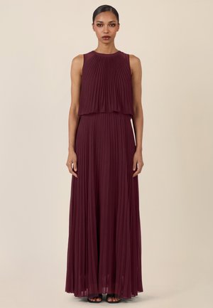 Kobieta o ciemnych włosach, ubrana w bezrękawnikową, plisowaną, bordową suknię maxi, stojąca na tle jednolitego beżowego tła.