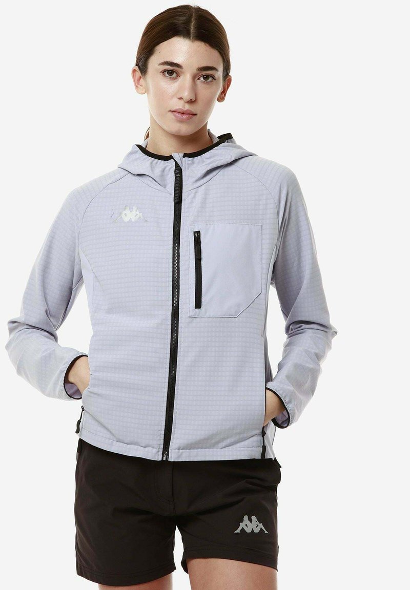 Giacca sportiva grigio chiaro con cappuccio, motivo a scacchi testurizzato, zip nera e tasca frontale. Abbinata a pantaloni corti neri con logo.