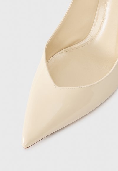 Pantof beige din piele lacuită cu toc înalt, cu vârful ascuțit și o tăietură în formă de V în partea superioară. Textură netedă și finisaj lucios.