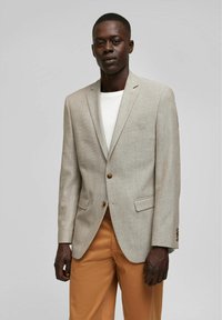 Beiger strukturierter Blazer mit einem Zwei-Knopf-Verschluss, Reverskragen und zwei vorderen Taschen. Getragen über einem weißen Hemd und kombiniert mit beigen Hosen.