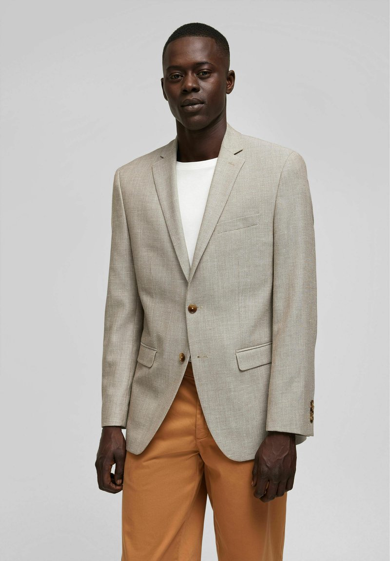 Beiger strukturierter Blazer mit einem Zwei-Knopf-Verschluss, Reverskragen und zwei vorderen Taschen. Getragen über einem weißen Hemd und kombiniert mit beigen Hosen.