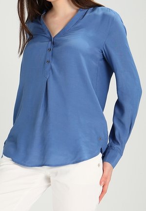 Vrouw draagt een blauwe blouse met lange mouwen en knopen, met een V-hals en witte broek, staand met één hand op de heup.