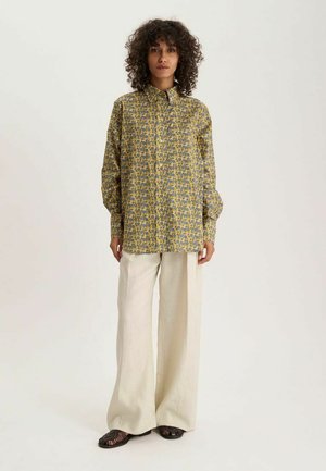 Femme aux cheveux bouclés portant une chemise jaune à motifs avec boutons, un pantalon large crème et des sandales noires tissées, debout sur fond blanc.