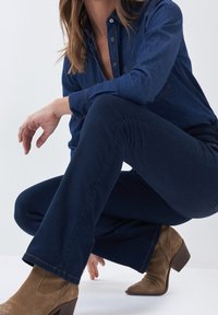 Chemise en denim bleu foncé, pantalon ajusté bleu, bottines en daim marron avec un talon empilé. Personne à genoux avec une main reposant sur la cuisse.