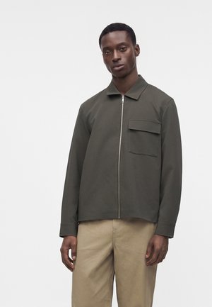 Homme portant une veste zippée vert olive avec une poche poitrine et un pantalon beige, debout devant un fond blanc uni.