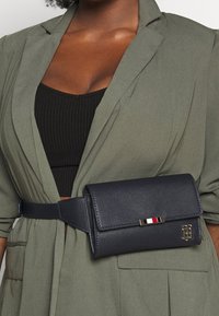 Námořnická kožená taška na opasek s texturovaným povrchem, zdobená zlatým kováním a malým pruhem v červené, bílé a modré barvě.