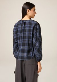 Czarna i niebieska koszula w kratę z długimi rękawami, marszczonym dekoltem i luźnym krojem; noszona z szarymi spodniami z wysokim stanem.