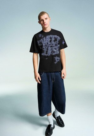 Sort bomulds T-shirt med lilla grafisk tekst, parret med oversized mørke denimshorts. Modellen har sorte sko og hvide sokker på.