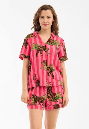 Kobieta ubrana w jasnoróżową koszulkę z krótkim rękawem i szorty z nadrukiem tygrysa i zielonej rośliny, stojąca na białym tle.