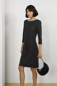 Abito nero con maniche a tre quarti, caratterizzato da arricciature sul lato e pannelli testurizzati. La modella tiene una borsa nera a forma curva.