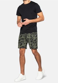 Uomo che indossa una maglietta nera, pantaloni corti mimetici verdi e neri, e sneakers bianche, in piedi su uno sfondo chiaro.
