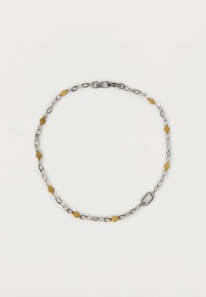 Srebrna verižica z enakomerno razporejenimi majhnimi zlatimi okroglimi poudarki in zaponko v obliki rakove kremplje na belem ozadju.