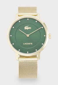 Montre verte ronde avec un bracelet en maille dorée, présentant un cadran texturé, des marqueurs d'heure en cristal et un logo de crocodile Lacoste à 12 heures.