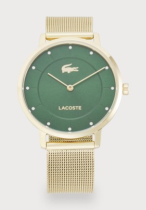 Montre verte ronde avec un bracelet en maille dorée, présentant un cadran texturé, des marqueurs d'heure en cristal et un logo de crocodile Lacoste à 12 heures.
