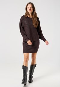 Ciemnobrązowy sweter z brokatowym splotem, zestawiony z dopasowaną spódnicą; modelka nosi czarne skórzane kozaki do kolan z akcentami klamrowymi.