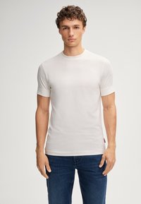 Junger Mann mit lockigem Haar, der ein tailliertes weißes Kurzarm-T-Shirt und dunkelblaue Jeans trägt und vor einem einfarbigen, hellen Hintergrund steht.