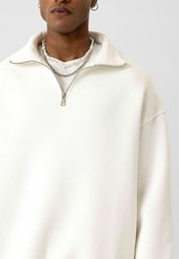 Witte zip-up sweater met een hoge kraag en een zachte textuur. Heeft een metalen rits en gelaagde kettingen voor extra detail.