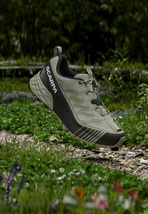 Lysegrå og sort Scarpa trail løbesko med hurtigsnørebånd, svævende over en grus- og græssti udendørs.