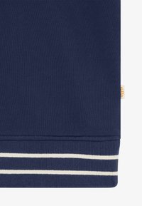 Granatowa tkanina z prążkowanym brzegiem, na którym znajdują się dwie białe poziome paski oraz mała beżowa metka z tkaniny po boku.