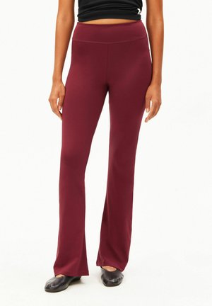 Frau trägt burgunderfarbene High-Waist Flare-Hose mit schwarzen Schuhen und kurzem schwarzem Oberteil, steht vor weißem Hintergrund.