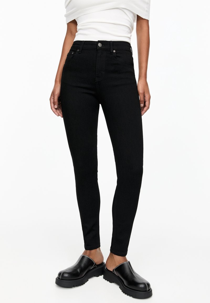 Jeans negros de cintura alta con un diseño ajustado, que presentan un cierre clásico de botón y cremallera. Combinados con zuecos negros y una blusa blanca.