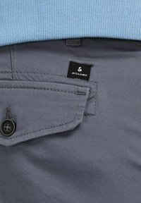 Grå tygbyxor med en framlapp ficka som har en svart knapp och en etikett som säger "JACK & JONES." Texturen är slät med en skräddarsydd design.