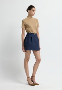Beige hoge hals top gecombineerd met een marineblauwe geruite minirok met een voorplooi en knoop. Model draagt bruine suède hakken.