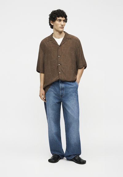 Άντρας με σγουρά μαλλιά που φοράει φαρδιά καφέ πουκάμισο με σχέδια πάνω σε λευκό μπλουζάκι, φαρδιά μπλε τζιν και μαύρα παπούτσια, στέκεται με τα χέρια μερικώς στις τσέπες.