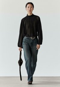 Sort bluse med langærmer og rynkede detaljer ved halsudskæringen, kombineret med blå jeans. Modellen holder et mørkebrunt bælte og en taske.