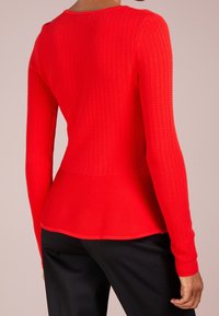 Rotes, gestricktes Langarmoberteil mit strukturiertem Muster, rundem Ausschnitt und ausgestellten Saum. Kombiniere es mit schwarzen Hosen, um den Farbkontrast zu betonen.