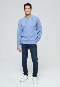 Homme portant un pull en maille torsadée bleu clair, un jean skinny noir et des baskets blanches, se tenant contre un fond gris clair.