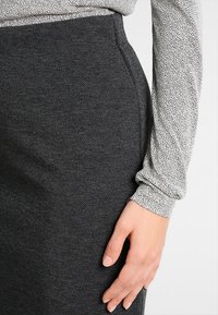 Jupe grise texturée au design ajusté, présentant un subtil motif en chevrons et des poches sur les côtés. Main reposant sur le tissu.