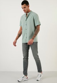 Camisa de botones de manga corta color verde menta con cuello mao, combinada con jeans grises desgastados y zapatillas blancas con acentos de color.