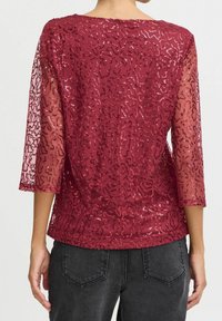 Kobieta w burgundowej, przetykanej cekinami przezroczystej bluzce z trzema ćwierć rękawami, zestawionej z ciemnoszarymi dżinsami, pokazana z tyłu.