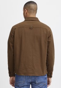 Camisa de algodón marrón con un cuello estructurado, detalles de botones en los puños y un lazo en la parte trasera. Se lleva con unos jeans de mezclilla azul.