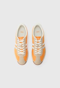 Orangene Sneaker mit beigen Akzenten, weißen Streifen und Schnürdesign. Weiches Obermaterial, Gummisohle und Markenlogo an der Seite.