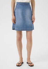 Jupe mini en denim bleu avec une coupe droite, présentant un ourlet cousu et des fentes latérales pour plus de confort. Portée avec des sandales noires à lanières.