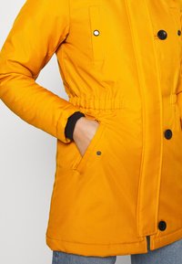 Personne portant un manteau jaune vif avec des boutons et des poches, main dans la poche, portant un jean bleu sur un fond clair uni.