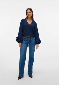 Blusa navy con pois testurizzati e scollatura a V, con maniche a campana arricciate. Abbinata a jeans dritti blu chiaro e scarpe marroni a punta.